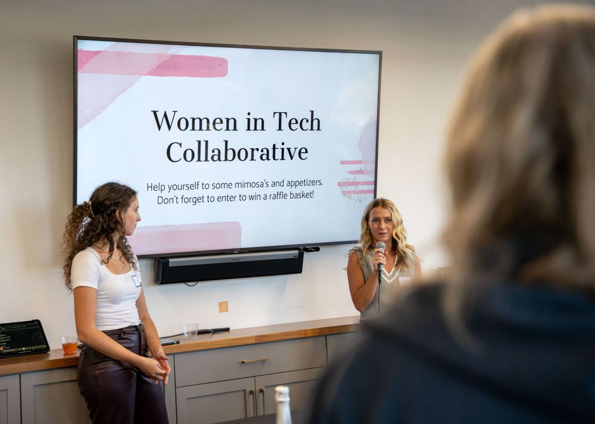 Two women speaking to a crowd with a screen that reads Women in Tech Collaborative behind them